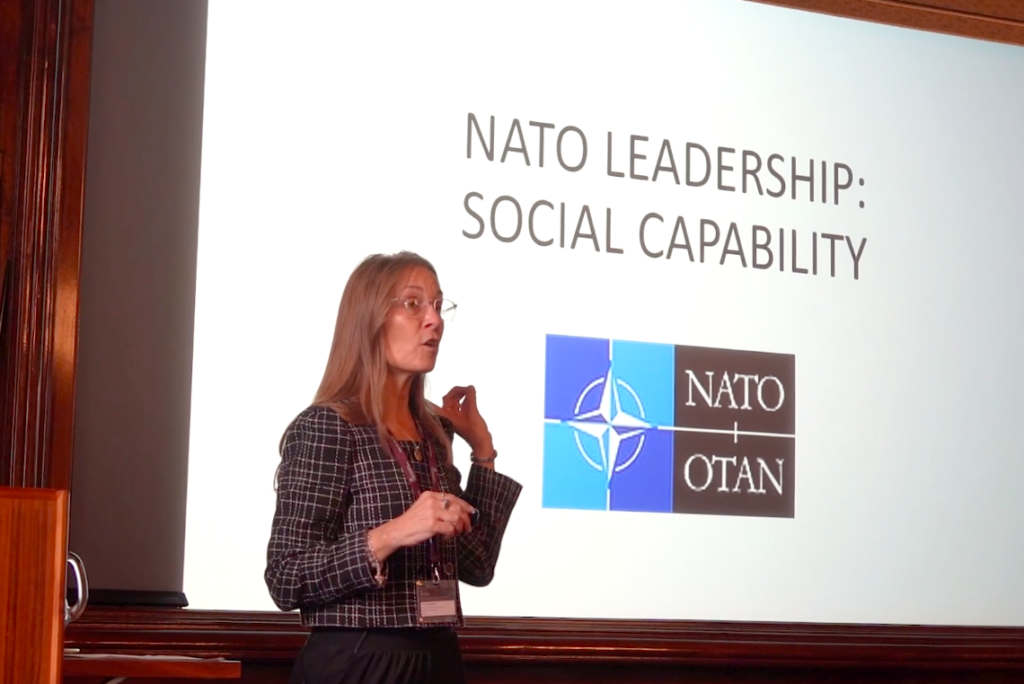 Dr. Laurie Mulvey in a grey suit jacket presenting in front of a slide that reads, "NATO Leadership: Social Capability" and includes the NATO/OTAN logo